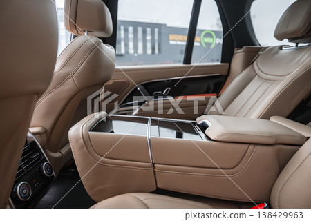Rear cabin of a luxury SUV shows tan leather captain chairs, a wide armrest with glossy controls, and vents, parked by an automotive service facility with large windows. Rear cabin of a luxury SUV shows tan leather captain chairs, a wide armrest with glossy controls, and vents, parked by an automotive service facility with large windows. 138429963