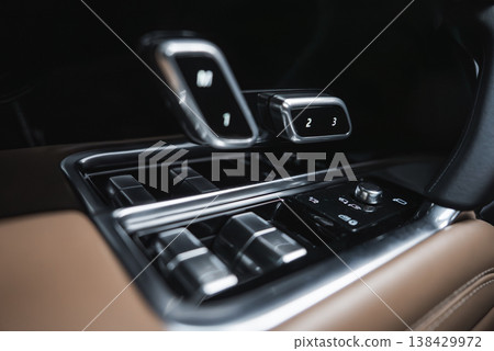 Close up of a luxury car console shows metallic toggles, rotary dial, glossy black panels, and dual digital gear selectors with M, 1, 2, 3 indicators under soft indoor light. 138429972