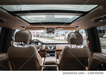 Wide angle view from second row shows tan leather seats, dual screens, and panoramic twin glass panels. Workshop or dealership lot with luxury cars is visible ahead. 138430046