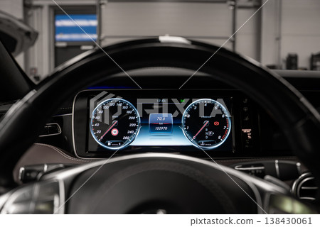 Driver's eye view shows twin gauges and steering wheel, speed and rpm flanking data screen, in a black dashboard within an upscale workshop under bright indoor light 138430061