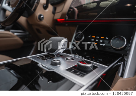 Close up of a luxury Range Rover Autobiography SUV console with gear selector, drive mode dial, and dual climate knobs under a glossy touchscreen, tan leather and red ambient light indoors 138430097