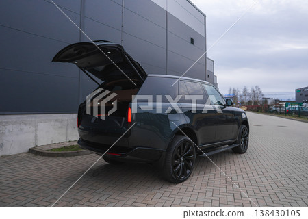 Black Range Rover Autobiography with tailgate open, vertical LED tail lights, black wheels, and tinted windows by gray metal warehouse in overcast daylight Black Range Rover Autobiography with tailgate open, vertical LED tail lights, black wheels, and tinted windows by gray metal warehouse in overcast daylight 138430105