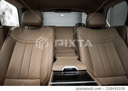 Tan leather rear seats with sculpted cushions and wide armrest with glossy controls and metallic trim, soft even lighting, view through rear window to indoor garage 138430109