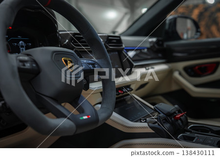 Stationary Lamborghini Urus SE close up shows suede steering wheel with bull emblem, tricolor badge, digital cluster, angular vents, touchscreen, and jet style selector. Stationary Lamborghini Urus SE close up shows suede steering wheel with bull emblem, tricolor badge, digital cluster, angular vents, touchscreen, and jet style selector. 138430111