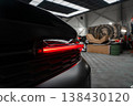 A close up shows the left rear LED taillight glowing on a black luxury SUV inside a high end workshop with skylights, tools, large wheels, and boxes in low key light. 138430120