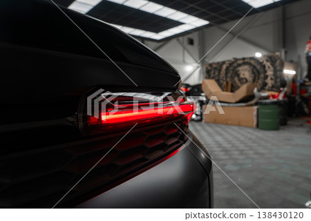 A close up shows the left rear LED taillight glowing on a black luxury SUV inside a high end workshop with skylights, tools, large wheels, and boxes in low key light. A close up shows the left rear LED taillight glowing on a black luxury SUV inside a high end workshop with skylights, tools, large wheels, and boxes in low key light. 138430120