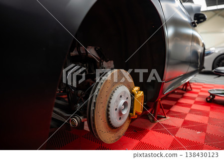 Close up of a luxury SUV on jack stands with front wheel off, showing drilled rotor and gold caliper on a red checkered floor in a professional service bay, low angle. 138430123