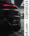 Matte black Lamborghini Urus rear quarter shows LED taillights, twin exhausts, and diffuser on a checkered garage floor, with cool overhead lighting suggesting service work. 138430124