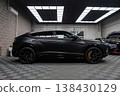 A matte black Lamborghini Urus SE sits in a modern workshop with checkered floor, grid lights, Y spoke wheels, and gold calipers as luxury cars undergo service nearby. 138430129