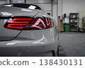 Close up shows silver Mercedes Benz Coupe AMG rear left quarter in a workshop. LED tail light glows red. Industrial shelving, green waste drum, and tools sit behind. 138430131