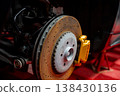 A close up shows a carbon ceramic disc, vented rotor, and gold caliper on a luxury sports SUV or coupe in a pro workshop, wheel off, parts lit with bright directional light. 138430136