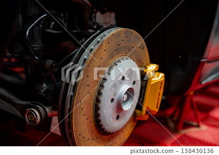 A close up shows a carbon ceramic disc, vented rotor, and gold caliper on a luxury sports SUV or coupe in a pro workshop, wheel off, parts lit with bright directional light. A close up shows a carbon ceramic disc, vented rotor, and gold caliper on a luxury sports SUV or coupe in a pro workshop, wheel off, parts lit with bright directional light. 138430136