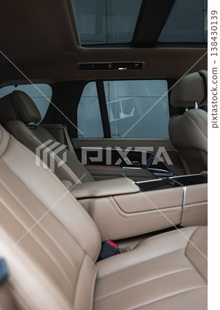 Close up of a luxury SUV rear cabin shows tan perforated seats, wide armrests, glossy black console, tinted windows, and a panoramic glass roof indoors in a showroom. 138430139