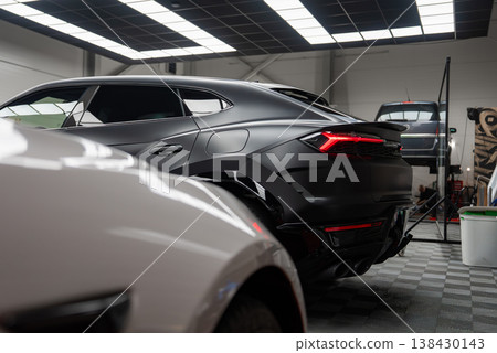 A matte black Lamborghini Urus stands in a lit workshop, sharp taillights visible. A Mercedes AMG and a new Range Rover are serviced, one on a lift, tools around. A matte black Lamborghini Urus stands in a lit workshop, sharp taillights visible. A Mercedes AMG and a new Range Rover are serviced, one on a lift, tools around. 138430143