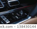 Close up of a premium car center console shows a glossy black panel, rotary dial, icon buttons, polished chrome, and tan leather inside a workshop setting. 138430144