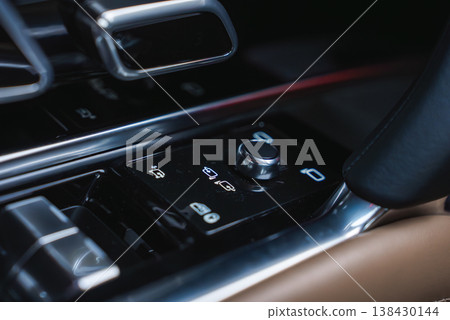 Close up of a premium car center console shows a glossy black panel, rotary dial, icon buttons, polished chrome, and tan leather inside a workshop setting. 138430144