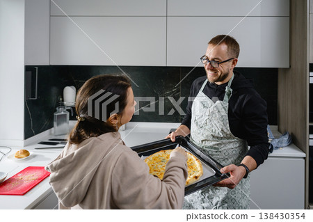 Happy couple holding baking tray with pizza and tasting it together in modern kitchen. Happy couple holding baking tray with pizza and tasting it together in modern kitchen. 138430354