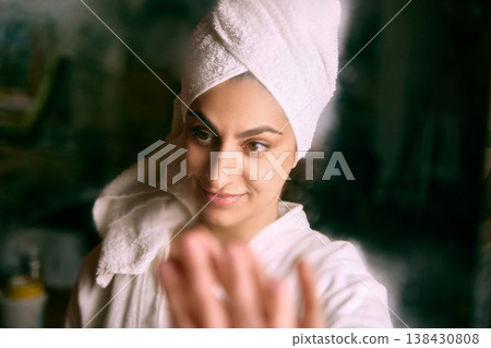 Woman in towel touching mirror during skincare routine. Woman in towel touching mirror during skincare routine. 138430808