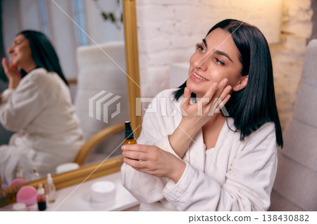 Woman applying facial cream looking in mirror at home 138430882