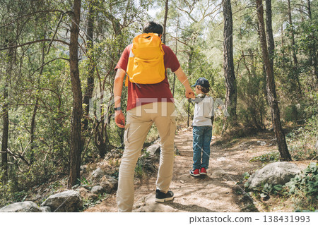 Rear view of tourists school boy and his dad walking a stone footpath in spring forest. Child boy and father wearing casual clothes and yellow backpack while hiking in summer greenwood leaf forest 138431993