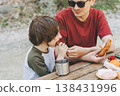 Close-up view of father hugs his school boy son on a family picnic. Child kid and his dad taking a rest and enjoying a picnic while hiking. Boy smiles and bites the bread donut on a picnic 138431996