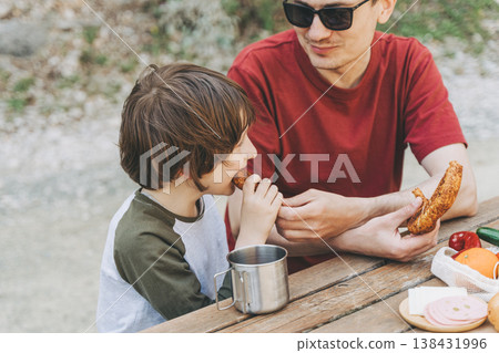 Close-up view of father hugs his school boy son on a family picnic. Child kid and his dad taking a rest and enjoying a picnic while hiking. Boy smiles and bites the bread donut on a picnic Close-up view of father hugs his school boy son on a family picnic. Child kid and his dad taking a rest and enjoying a picnic while hiking. Boy smiles and bites the bread donut on a picnic 138431996