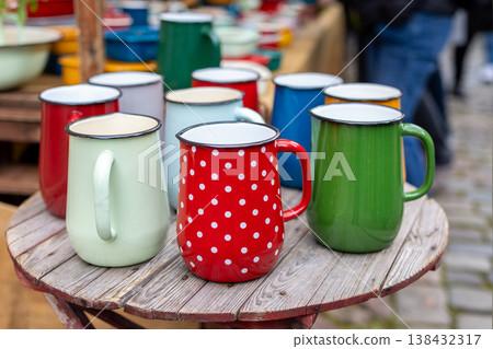Colorful enamel mugs arranged rustic wooden table outdoor market stall. Vintage metal cups bright colors polka pattern cheerful display classic camping style kitchenware popular coffee tea picnic use Colorful enamel mugs arranged rustic wooden table outdoor market stall. Vintage metal cups bright colors polka pattern cheerful display classic camping style kitchenware popular coffee tea picnic use 138432317