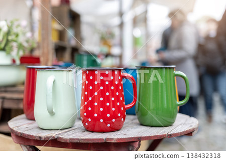 Colorful enamel mugs arranged rustic wooden table outdoor market stall. Vintage metal cups bright colors polka pattern cheerful display classic camping style kitchenware popular coffee tea picnic use Colorful enamel mugs arranged rustic wooden table outdoor market stall. Vintage metal cups bright colors polka pattern cheerful display classic camping style kitchenware popular coffee tea picnic use 138432318