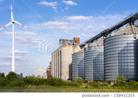 Large agricultural grain silo conveyor system industrial storage facility area wind turbine blue sky. Rural industry infrastructure combine crop storage logistic operation renewable energy production 138432355