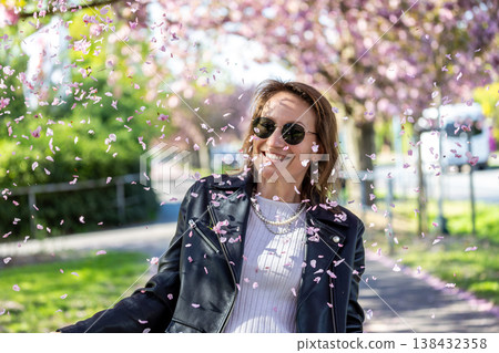 Smiling woman standing under blooming sakura trees pink petals fall around her in sunny spring park. Joyful lifestyle moment during cherry blossom season soft floral atmosphere bright daylight 138432358