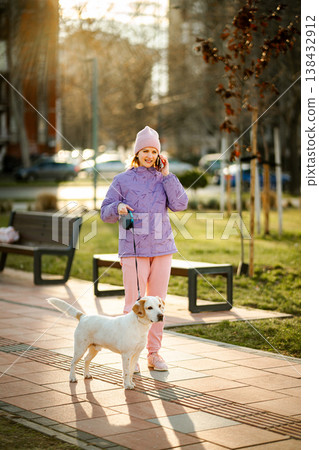 Woman talking on smartphone while walking dog in park 138432912