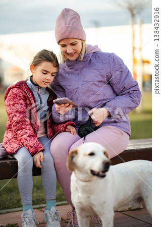 Mother and daughter using smartphone with dog in park 138432916