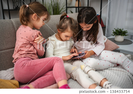 Three little girls sharing a mobile device on sofa Three little girls sharing a mobile device on sofa 138433754