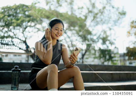 Smiling woman resting and listening to music on smartphone outdoor workout break in park Smiling woman resting and listening to music on smartphone outdoor workout break in park 138435091