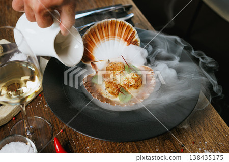 Grilled scallops served in a shell with avocado mousse and microgreens. Elegant seafood dish presented with dry ice smoke. Gourmet cuisine in a fine dining restaurant Grilled scallops served in a shell with avocado mousse and microgreens. Elegant seafood dish presented with dry ice smoke. Gourmet cuisine in a fine dining restaurant 138435175