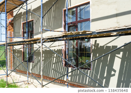 Exterior facade finishing, plastering stage, scaffolding setup, wall leveling, surface preparation for decorative render, house construction works. Exterior facade finishing, plastering stage, scaffolding setup, wall leveling, surface preparation for decorative render, house construction works. 138435648