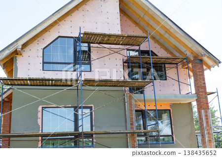House under facade plastering, exterior scaffolding for render application, insulation layer, surface leveling, construction wall finish, decorative coating. House under facade plastering, exterior scaffolding for render application, insulation layer, surface leveling, construction wall finish, decorative coating. 138435652