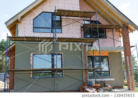 Render application to facade, scaffolding at exterior finishing stage, mesh-reinforced waterproof wall, thermal insulation, smooth surface, modern house build. 138435653
