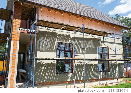 Plaster coating on facade insulation, external wall scaffolded for finishing, construction phase, smoothing render, surface preparation, house renovation works. Plaster coating on facade insulation, external wall scaffolded for finishing, construction phase, smoothing render, surface preparation, house renovation works. 138435654