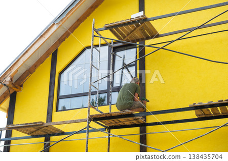 Applying finish paint to facade plaster, house exterior wall, renovation project, yellow color, wood decorative strips, maintenance step, final wall protection. Applying finish paint to facade plaster, house exterior wall, renovation project, yellow color, wood decorative strips, maintenance step, final wall protection. 138435704
