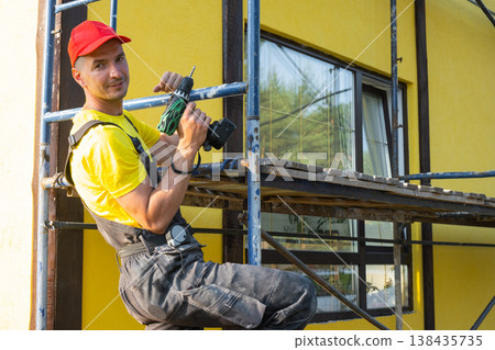 Builder in red cap with screwdriver on scaffolding, house facade, home repair, instrument kit, handyman visiting for at-home craft, maintenance and improvement. Builder in red cap with screwdriver on scaffolding, house facade, home repair, instrument kit, handyman visiting for at-home craft, maintenance and improvement. 138435735