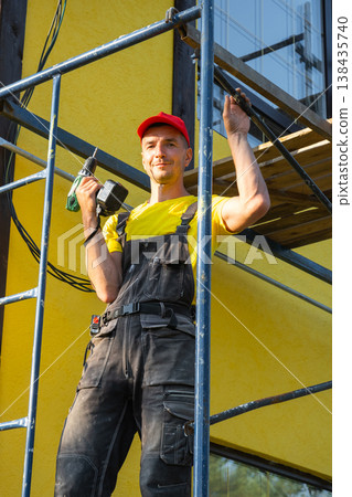 Repairman in overalls, on scaffold with paint roller, facade painting, consulting for exterior house renovation, on-site service, handyman tool kit. Repairman in overalls, on scaffold with paint roller, facade painting, consulting for exterior house renovation, on-site service, handyman tool kit. 138435740