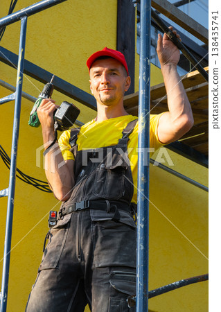 Handyman holding paint roller and power tool, scaffolding for home repairs, exterior wall renovation, full service call, craftsman visiting for consulting. Handyman holding paint roller and power tool, scaffolding for home repairs, exterior wall renovation, full service call, craftsman visiting for consulting. 138435741