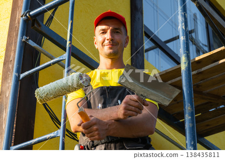 Portrait of young builder with large paint roller, facade renovation, scaffold work, maintenance, home visit, full handyman service, tools for repairs. Portrait of young builder with large paint roller, facade renovation, scaffold work, maintenance, home visit, full handyman service, tools for repairs. 138435811