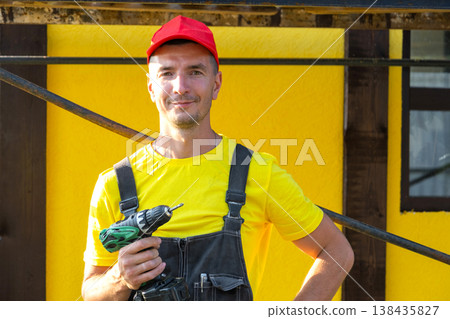 Builder portrait holding drill, standing near house under construction, repair services, private handyman business, tools, DIY home improvement skills. Builder portrait holding drill, standing near house under construction, repair services, private handyman business, tools, DIY home improvement skills. 138435827