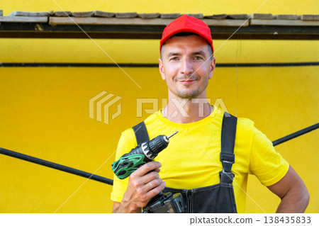 Builder with cordless drill and toolkit, standing at house under construction, at-home repairs, professional contractor business, DIY improvement skills. Builder with cordless drill and toolkit, standing at house under construction, at-home repairs, professional contractor business, DIY improvement skills. 138435833