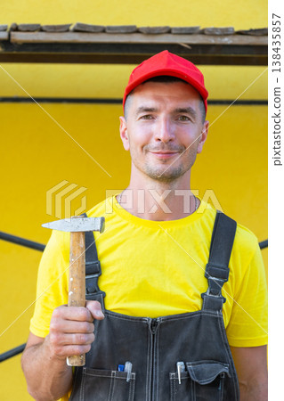 Portrait of builder in red cap holding hammer, private handyman business, home repairs, carpentry, on-site DIY construction, home building expert. Portrait of builder in red cap holding hammer, private handyman business, home repairs, carpentry, on-site DIY construction, home building expert. 138435857