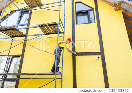 Master attaches vertical wooden decorative elements to facade, half-timbered style, exterior house finish, building improvement, private project work. Master attaches vertical wooden decorative elements to facade, half-timbered style, exterior house finish, building improvement, private project work. 138435892