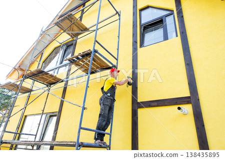Craftsman in red cap fixes decorative wood on high facade, external home finish, half-timber construction, cladding improvement, mounting work at height. Craftsman in red cap fixes decorative wood on high facade, external home finish, half-timber construction, cladding improvement, mounting work at height. 138435895