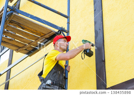 Master fixes timber element to house wall, half-timber (fachwerk) design, decorative cladding, home exterior DIY improvement, private renovation work. Master fixes timber element to house wall, half-timber (fachwerk) design, decorative cladding, home exterior DIY improvement, private renovation work. 138435898